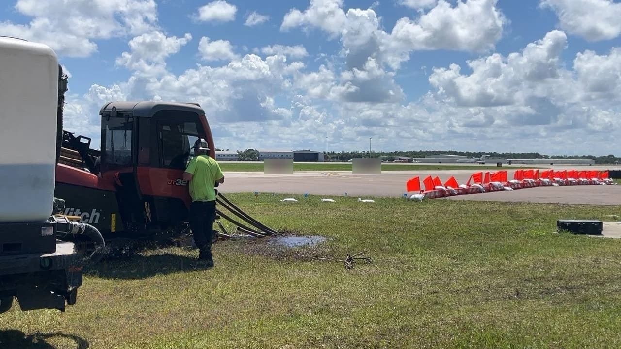 HDD contractor crew member at a Ditch Witch JT32 rig on an airfield, lines running from the rig to a pool of drilling fluid on the grass