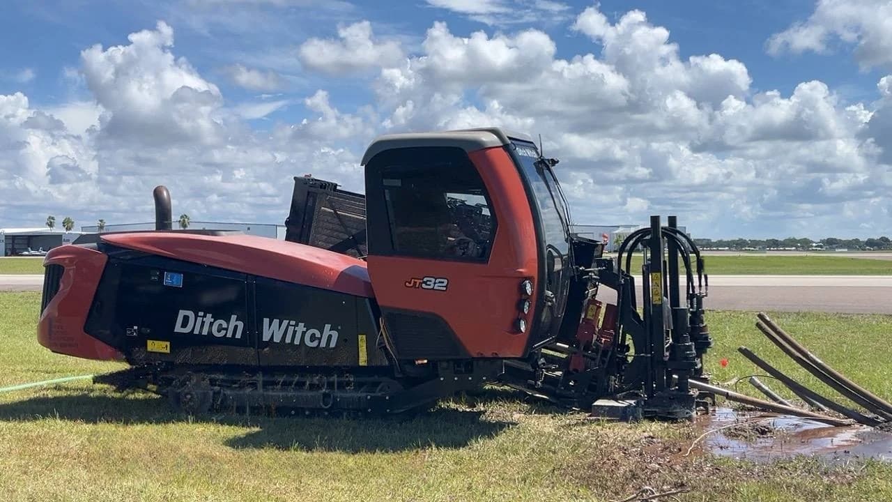 Ditch Witch JT32 horizontal directional drilling rig set up on grass beside an airfield, drill rod laid out next to the rig