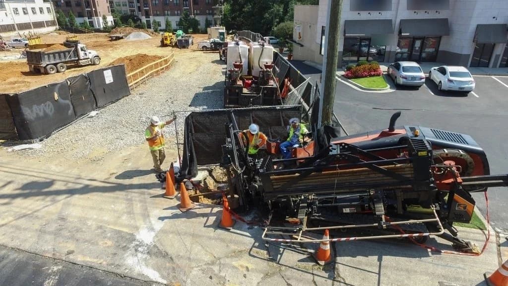 Aerial view of a directional drill working inside a fenced work zone on a paved commercial site, cones and crew around the bore pit, a trenchless pipe installation with no open trench