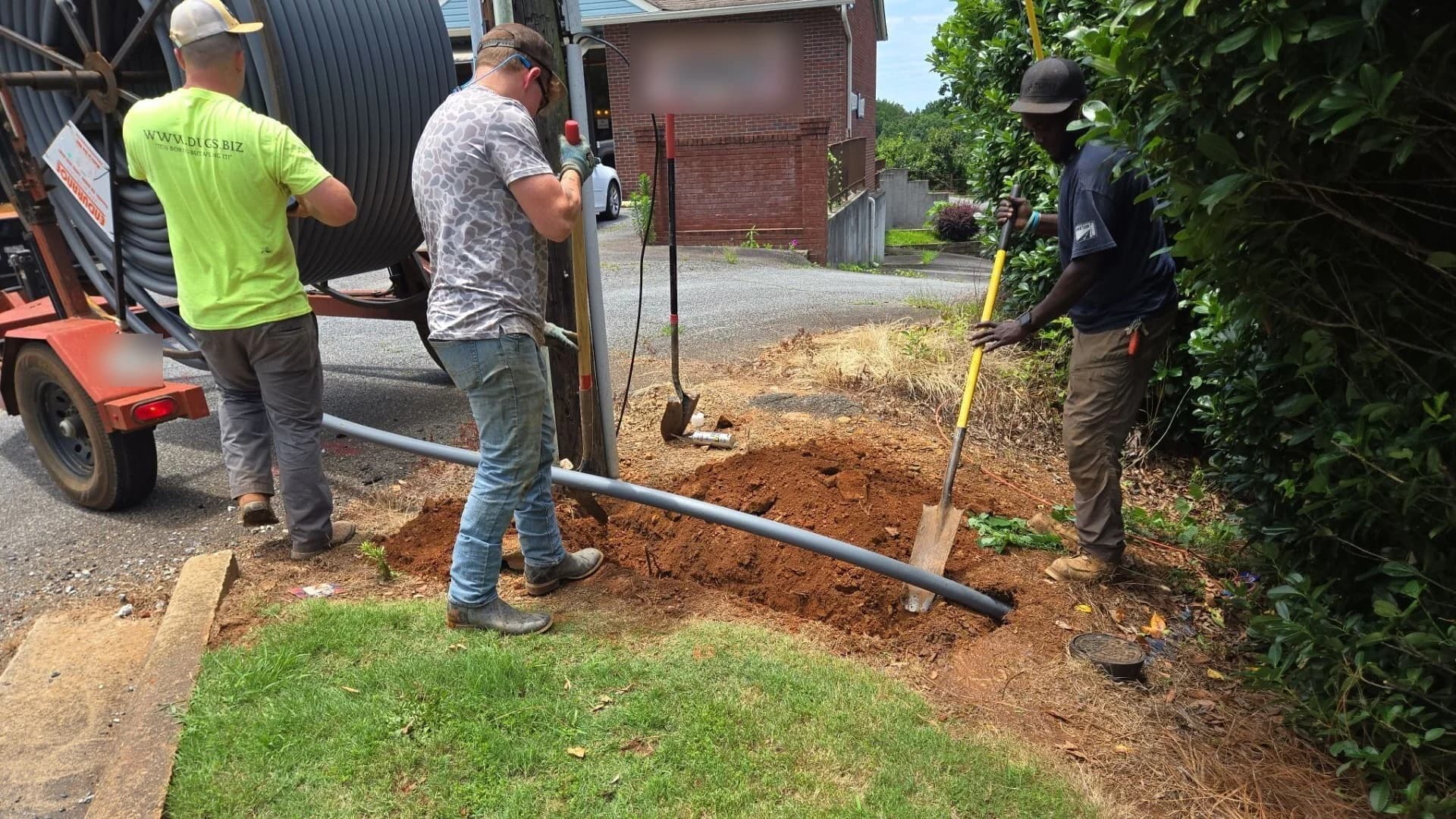 Crew unspooling gray HDPE conduit from a reel trailer while a worker hand digs the entry pit for a directional boring job beside a driveway